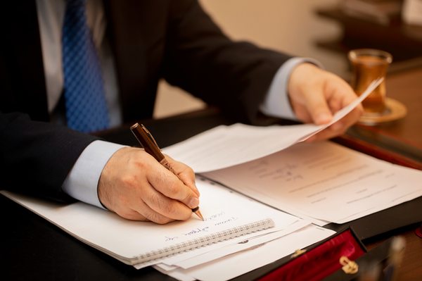 A close-up of a businessman’s hands writing on a notepad while reviewing documents on a desk, conveying professionalism and focus.